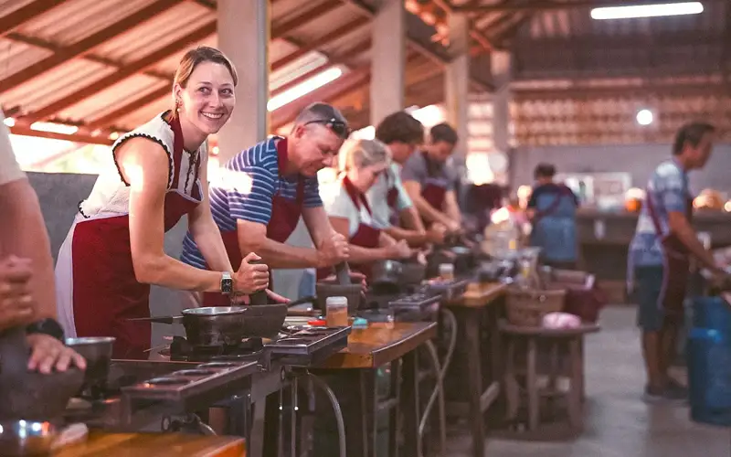 Travelers smiling and cooking at individual wok stations during a group Thai cooking class in Chiang Mai. Travelers smiling and cooking at individual wok stations during a group Thai cooking class in Chiang Mai.