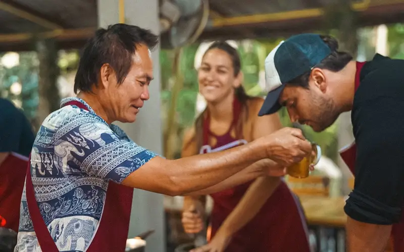 Local Thai instructor showing a student how to taste and adjust seasoning during a hands-on cooking class. Local Thai instructor showing a student how to taste and adjust seasoning during a hands-on cooking class.